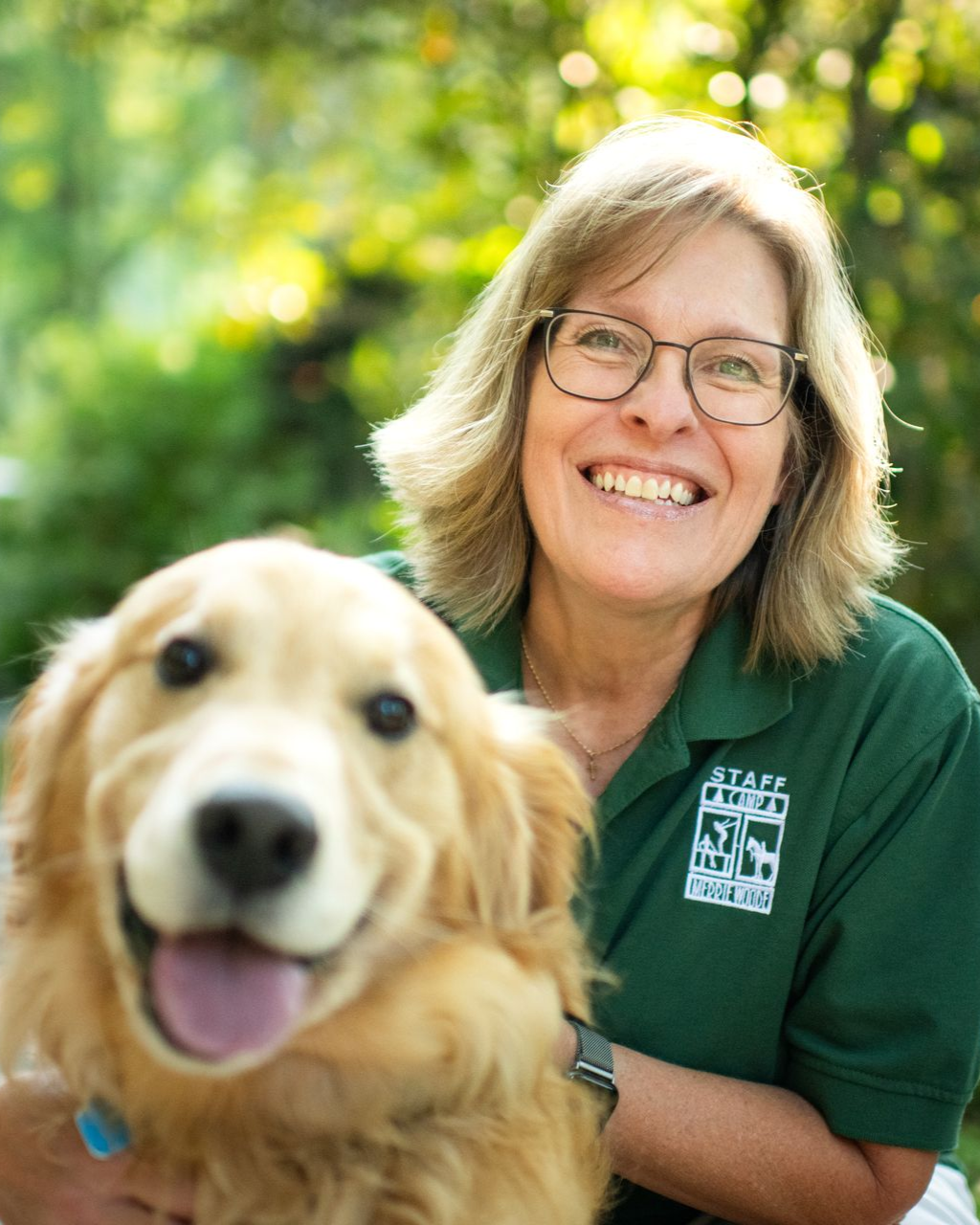 A smiling person wearing a green polo shirt poses outdoors next to a happy golden retriever.