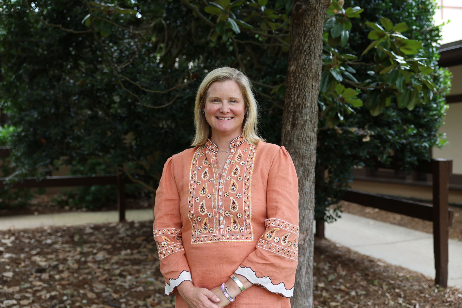 A person with blonde hair smiling, wearing a white eyelet shirt under a blue denim jacket, outdoors against green trees.