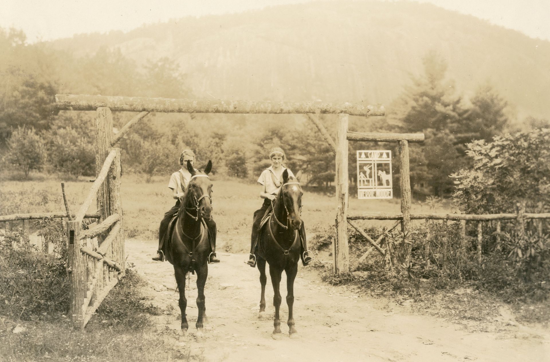Two people on horseback ride through a rustic wooden gateway into a clearing with trees and a mountain in the background.