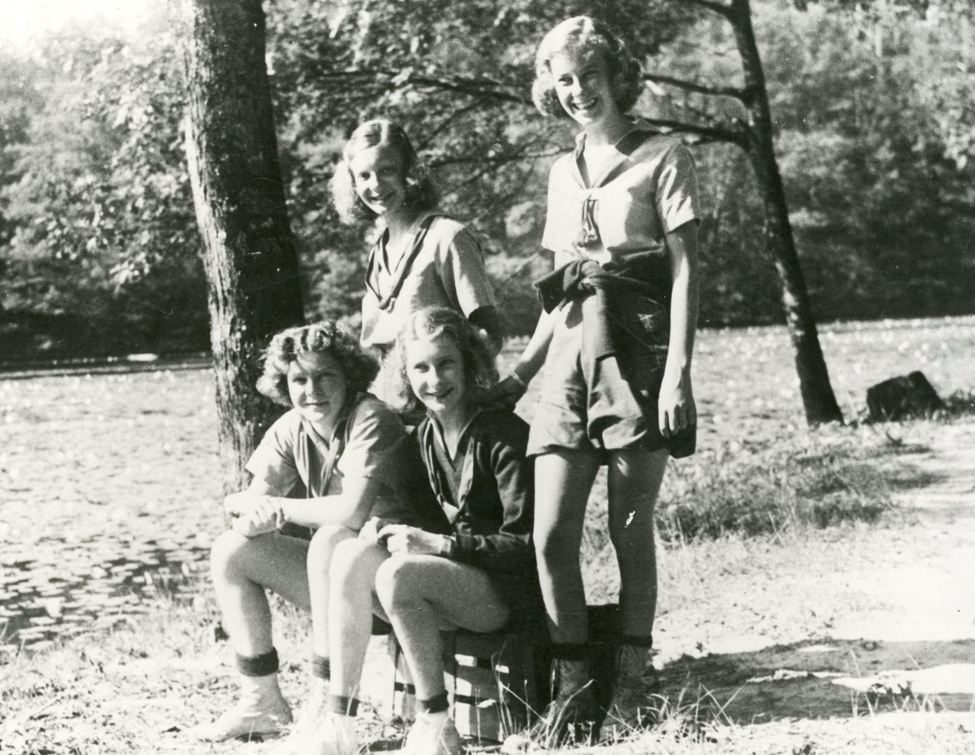 Four individuals pose together in an outdoor, wooded setting near a grassy area and trees, wearing casual warm-weather attire.