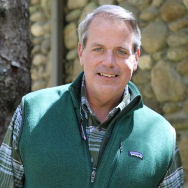A person wearing a green fleece vest over a plaid shirt, smiling in front of a stone wall and a tree.