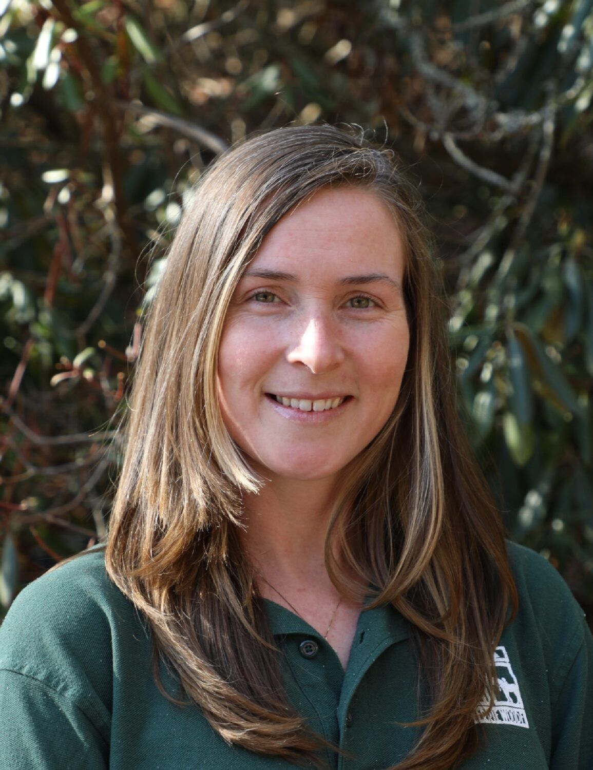 A smiling person with long brown hair wearing a dark green polo shirt, standing in front of out-of-focus foliage.