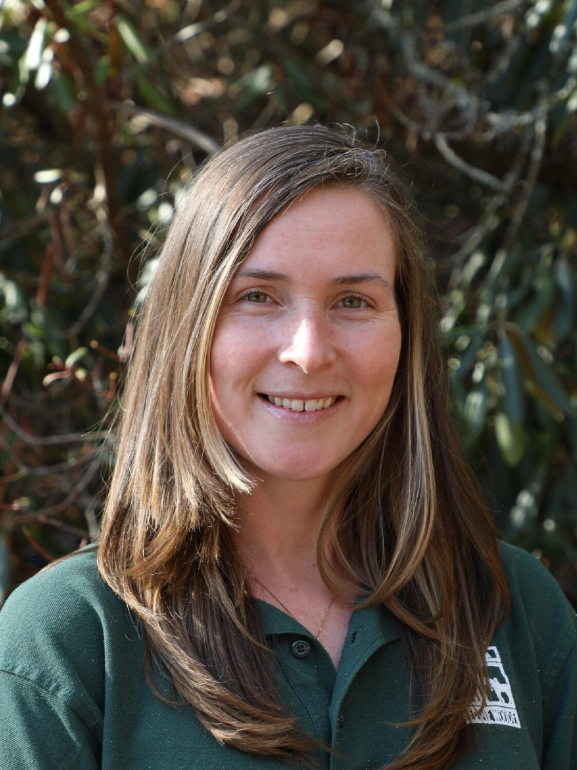 A smiling person with long brown hair wearing a dark green polo shirt, standing in front of out-of-focus foliage.