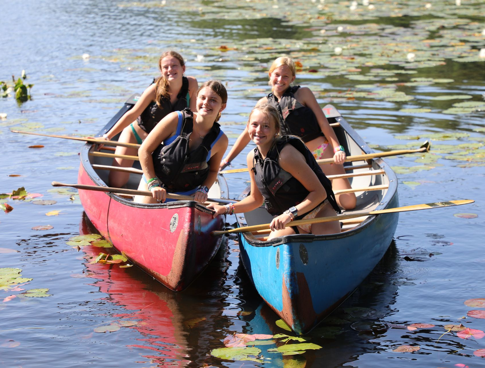 Four people in life vests paddle a red canoe and a blue canoe through lily pads on a sunny day, smiling at the camera.