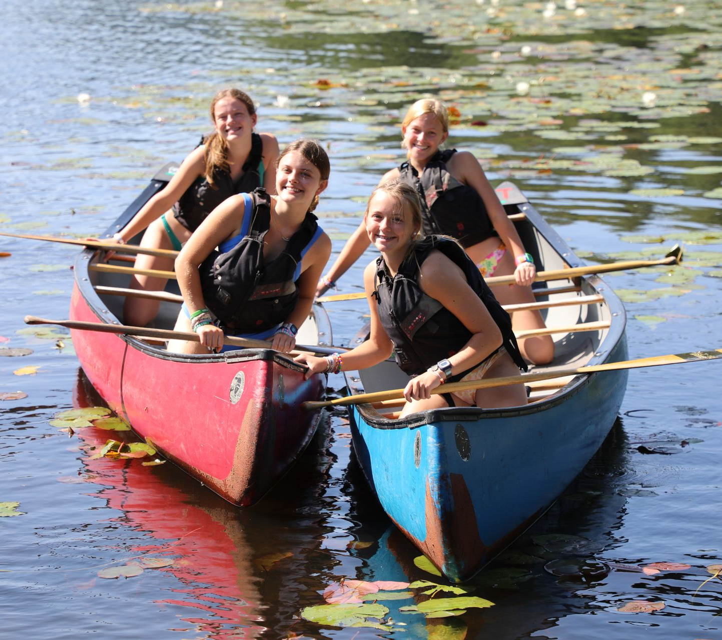 Four people in life vests smile while paddling in two colorful canoes on a calm, lily-pad-filled lake.