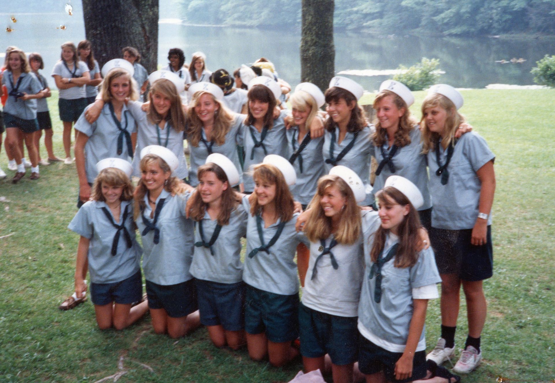 A group of people in matching uniforms and white hats pose outdoors by a lake, some standing and some kneeling.