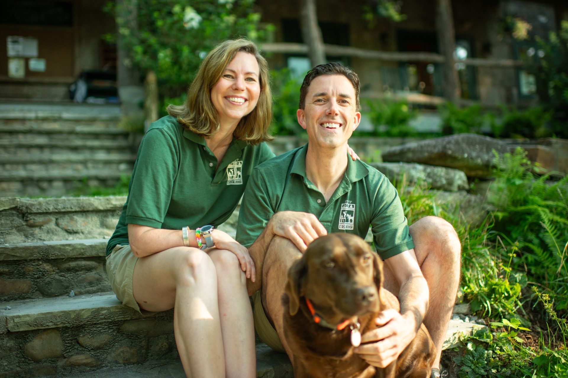 A smiling couple wearing matching green polo shirts sit on stone steps outdoors with a brown dog.