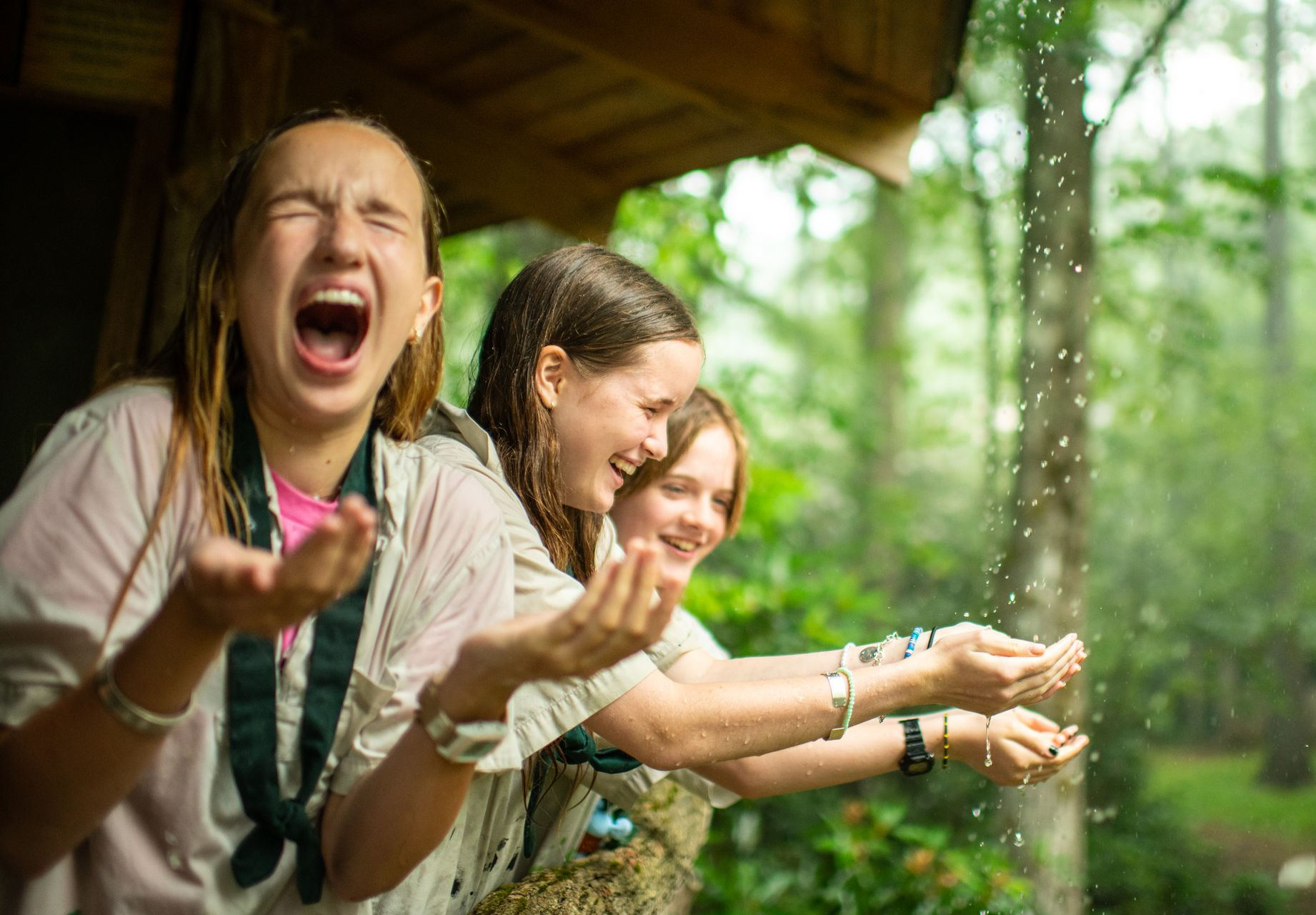 Three people stand on a porch in the rain, hands outstretched, laughing with joy amidst the forest setting.