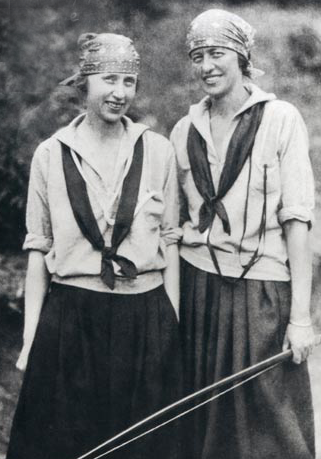 Two individuals wearing headscarves, collared shirts, and dark skirts stand side-by-side smiling in a natural setting.