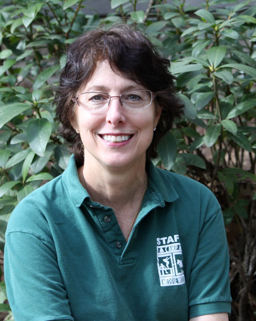 A person wearing a green polo shirt with a logo, smiling in front of a green leafy bush.