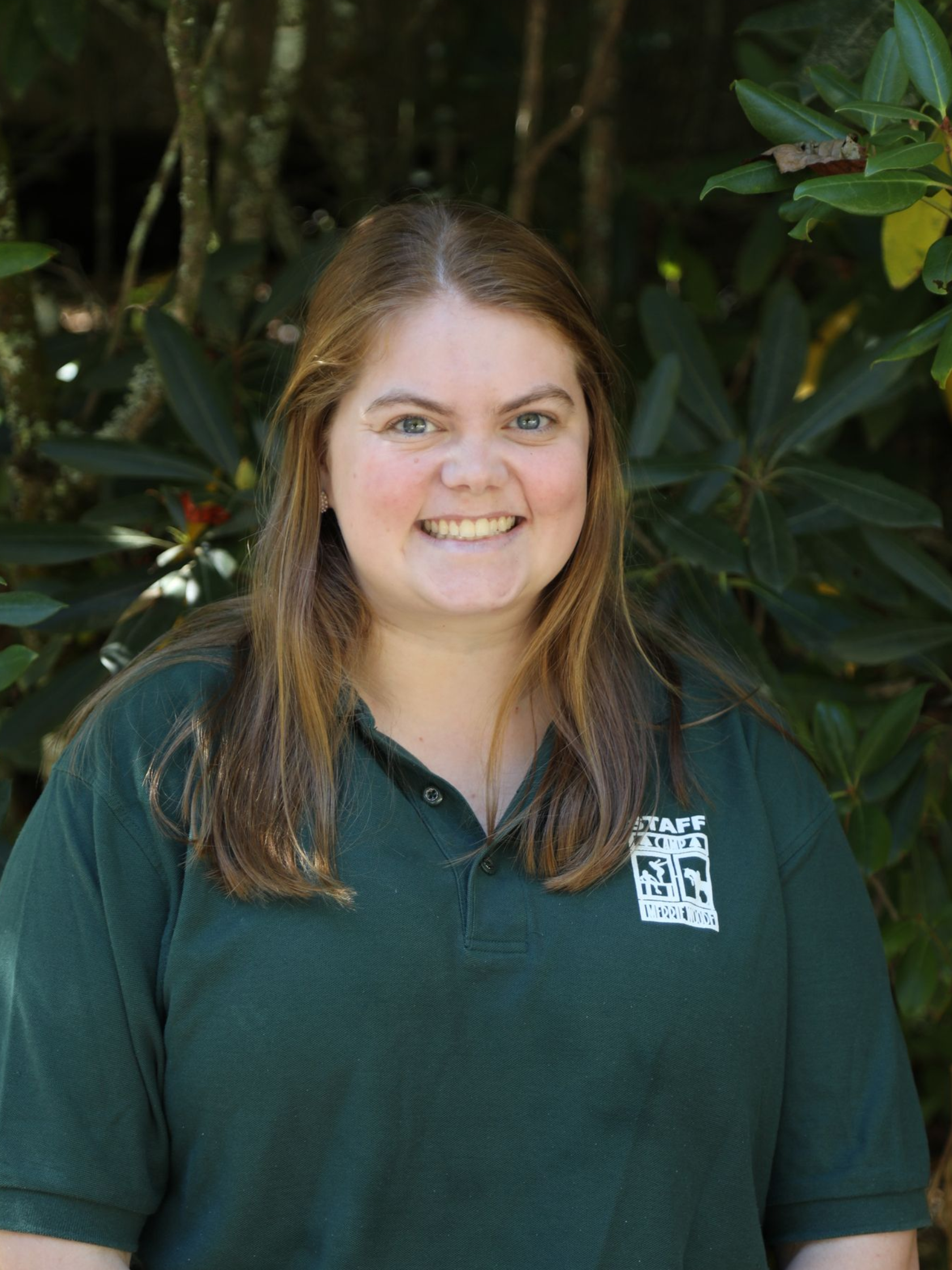 A smiling individual with long blonde hair, wearing a dark green polo shirt with a white logo, posed against leafy greenery.