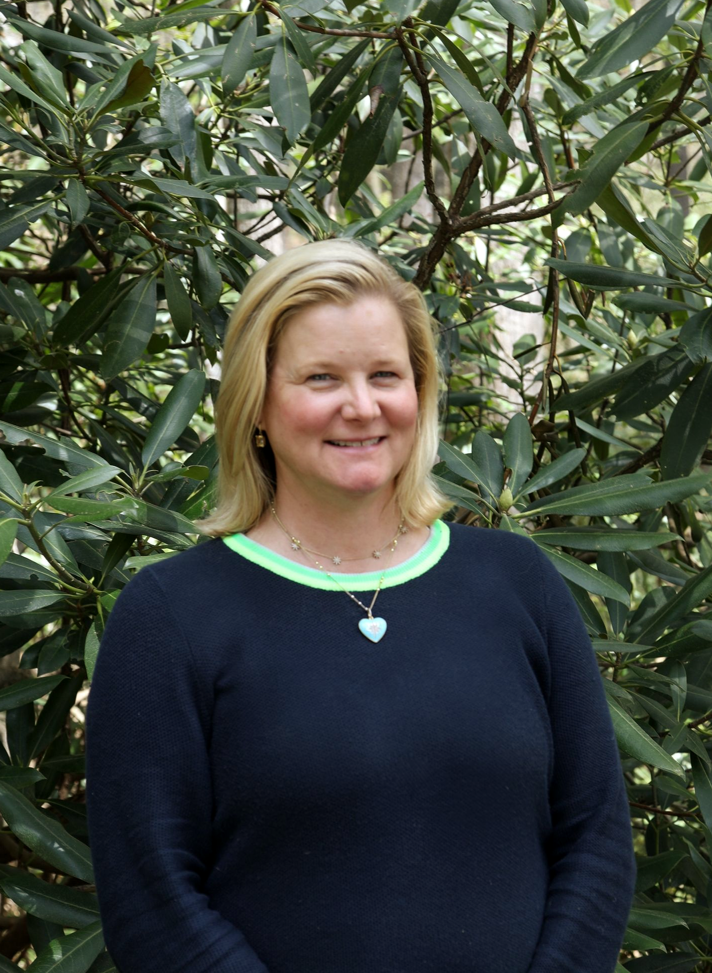 A person with blonde hair smiling, wearing a white eyelet shirt under a blue denim jacket, outdoors against green trees.