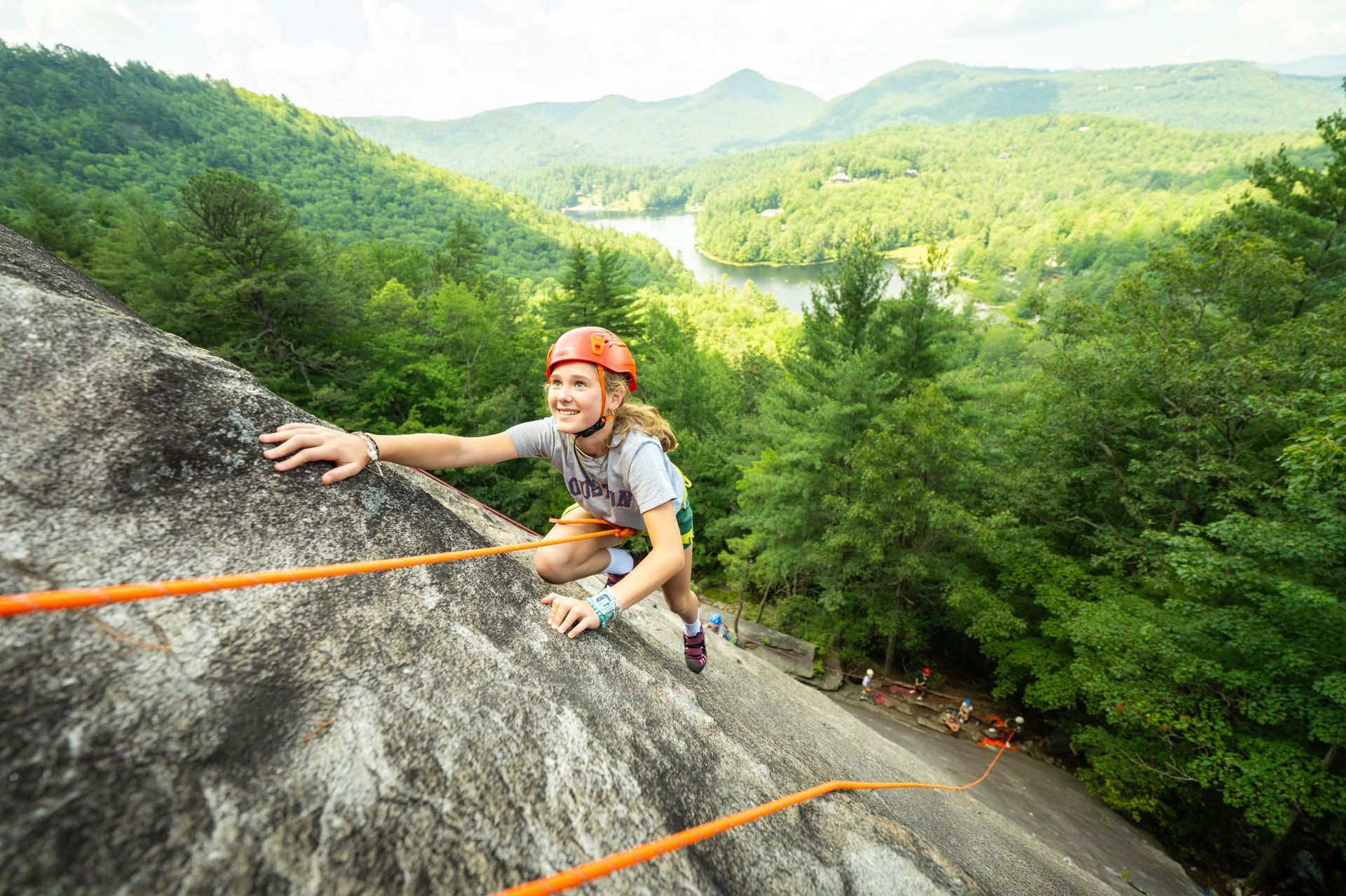 A climber in a helmet scales a rocky cliff overlooking a lush green valley with a river.