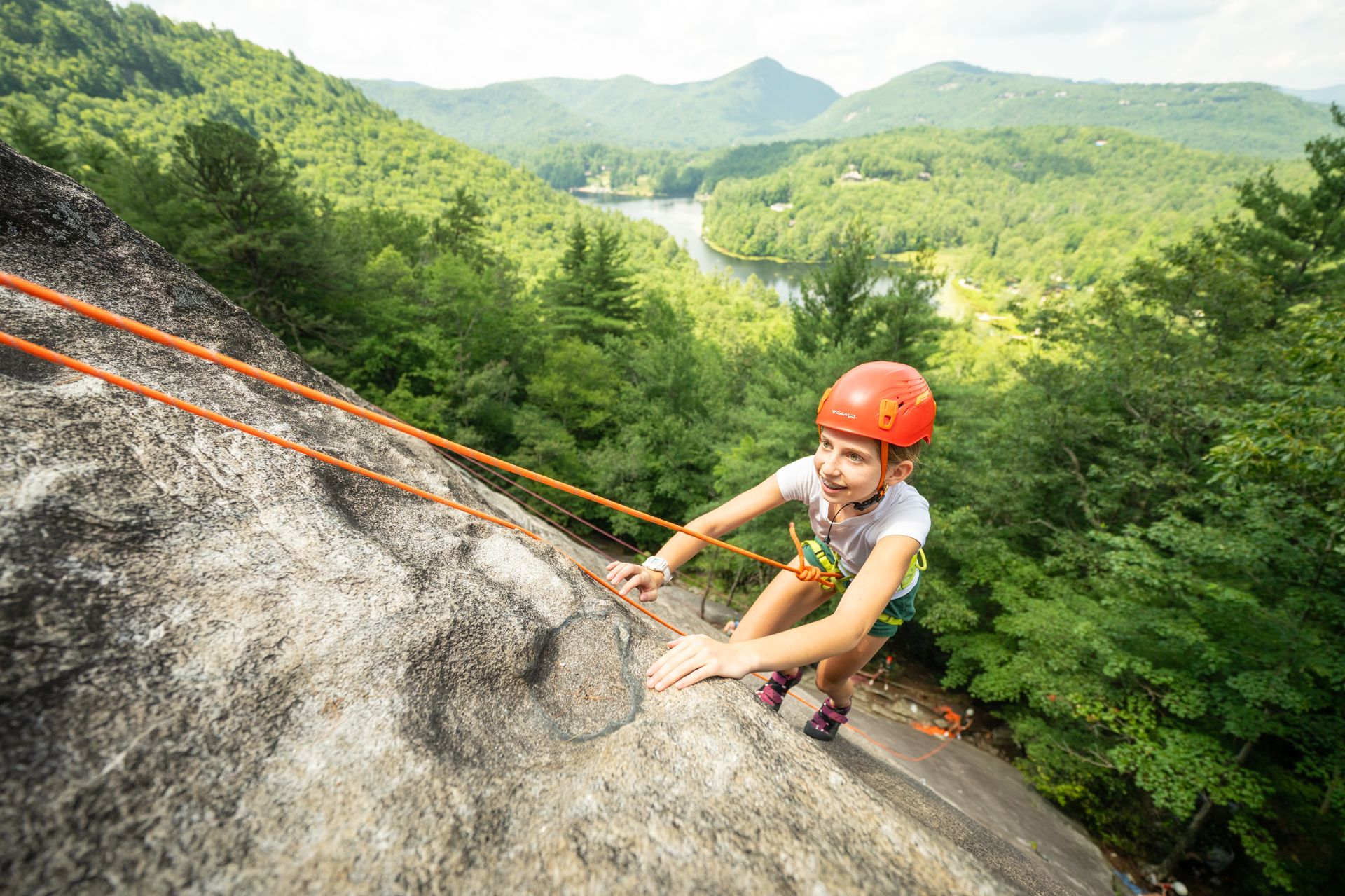 A climber wearing a bright orange helmet ascends a steep rock face overlooking a forested valley with a lake.
