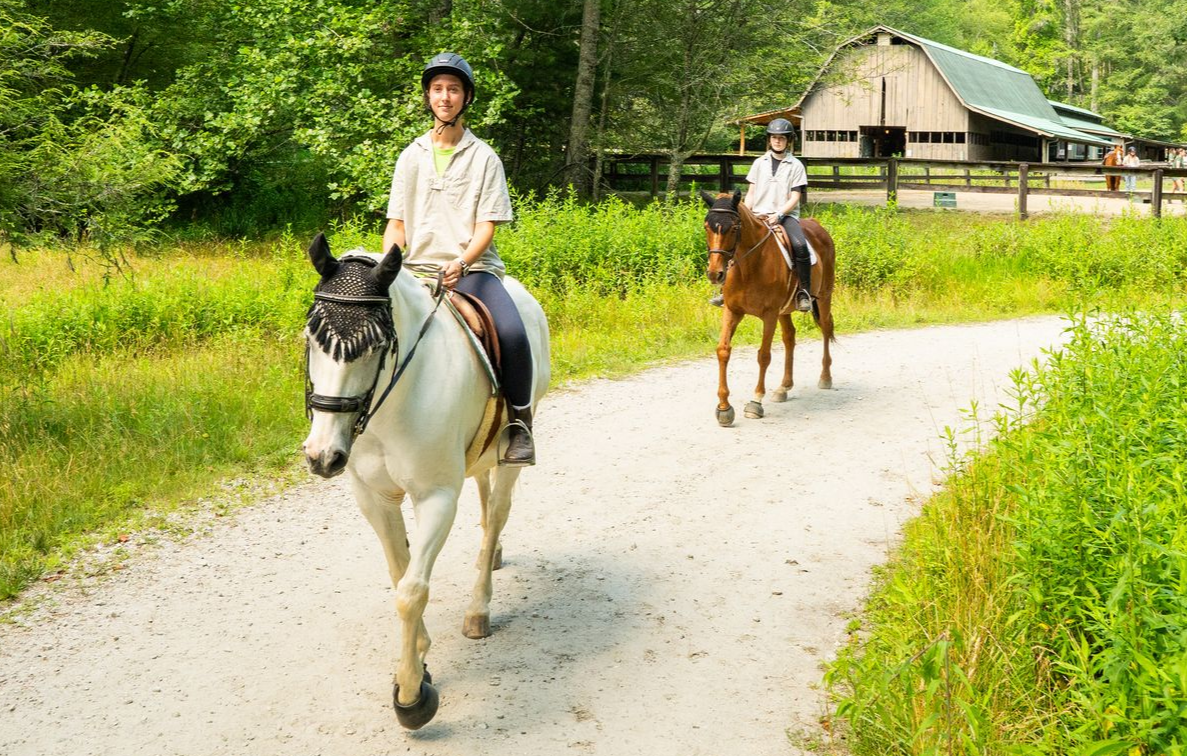Two riders on horseback travel along a gravel path toward a wooden barn on a sunny day surrounded by lush green foliage.