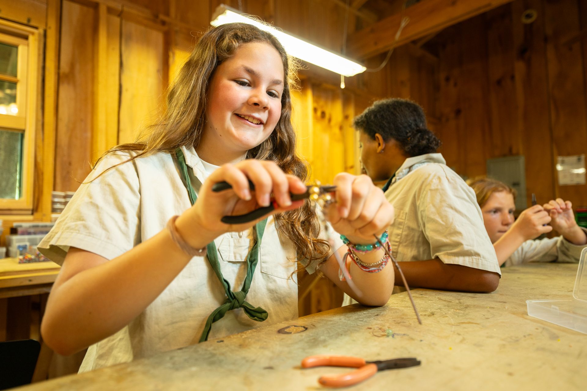 Three people in a wooden workshop craft jewelry with wire and pliers, focusing intently on their projects.