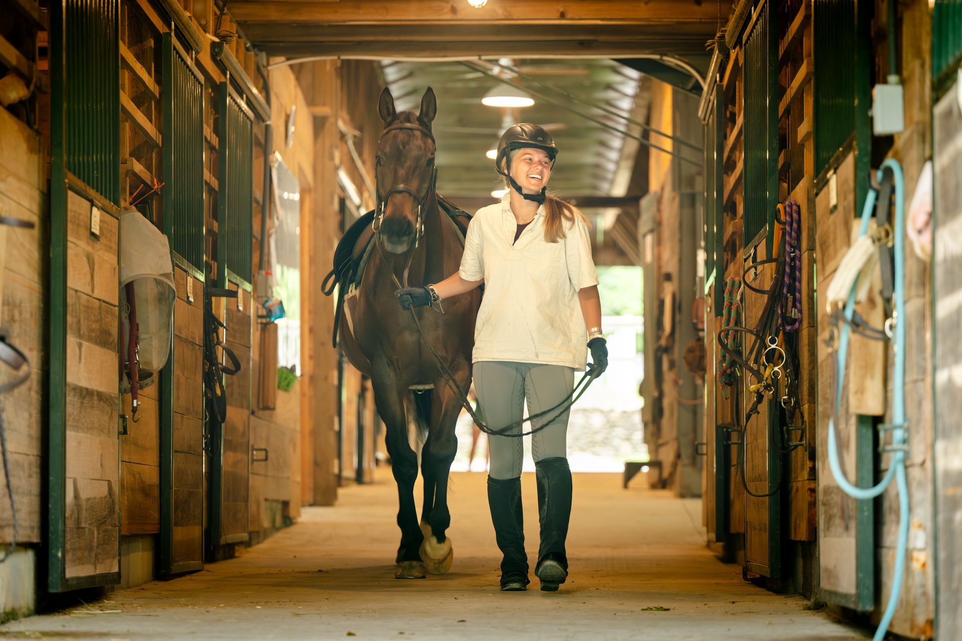 A person in a riding helmet and light-colored shirt leads a saddled brown horse down the aisle of a wooden horse stable.
