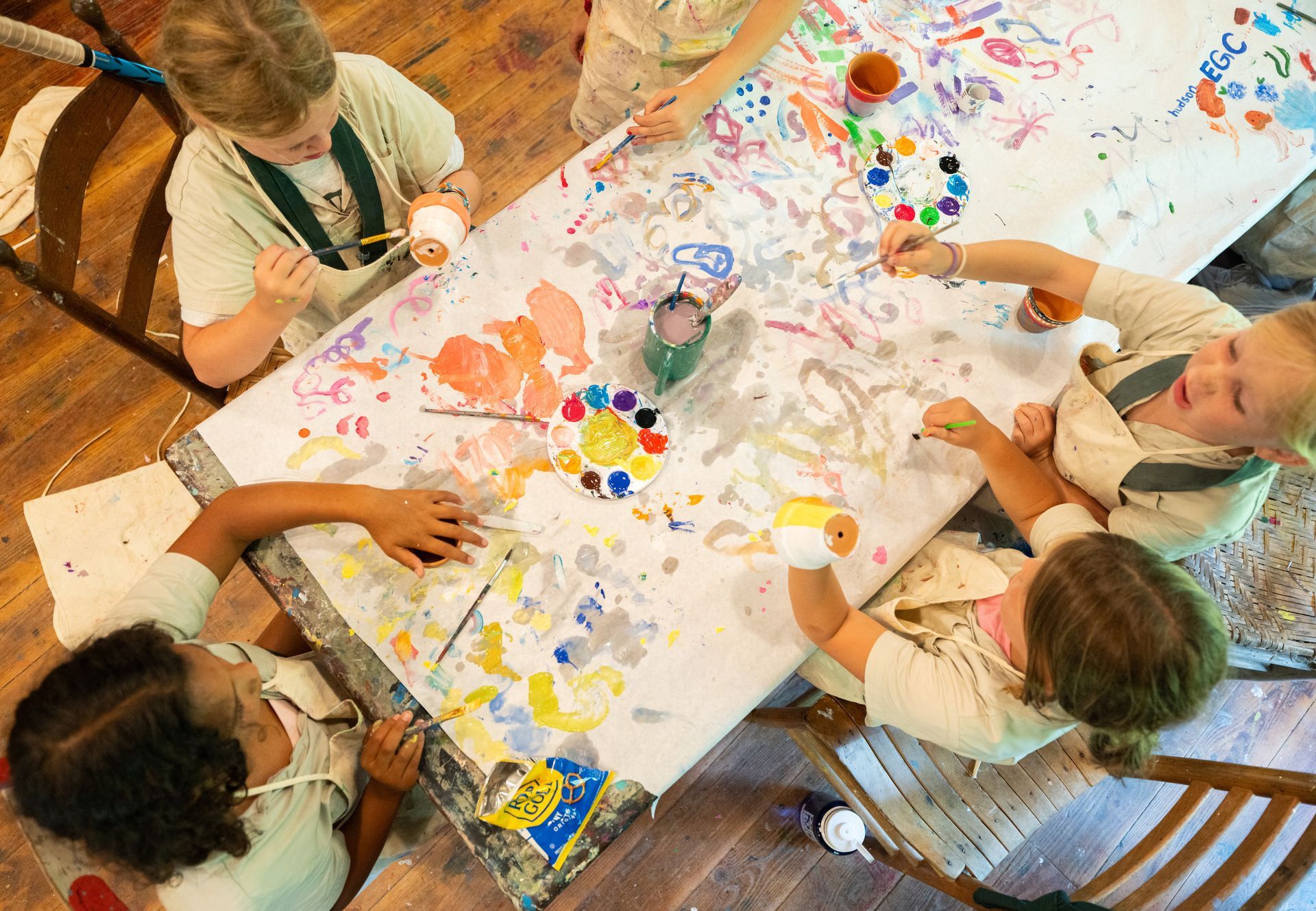 Four people painting on a large paper-covered table together in an art studio.