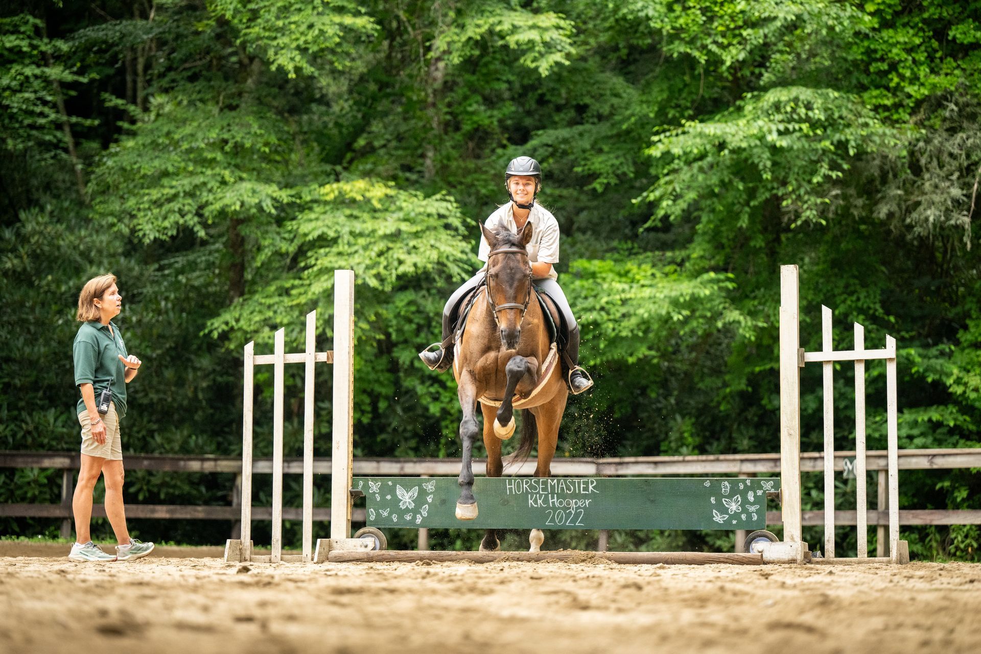 A rider on a brown horse jumps over a green obstacle in a wooded outdoor arena as an instructor watches nearby.