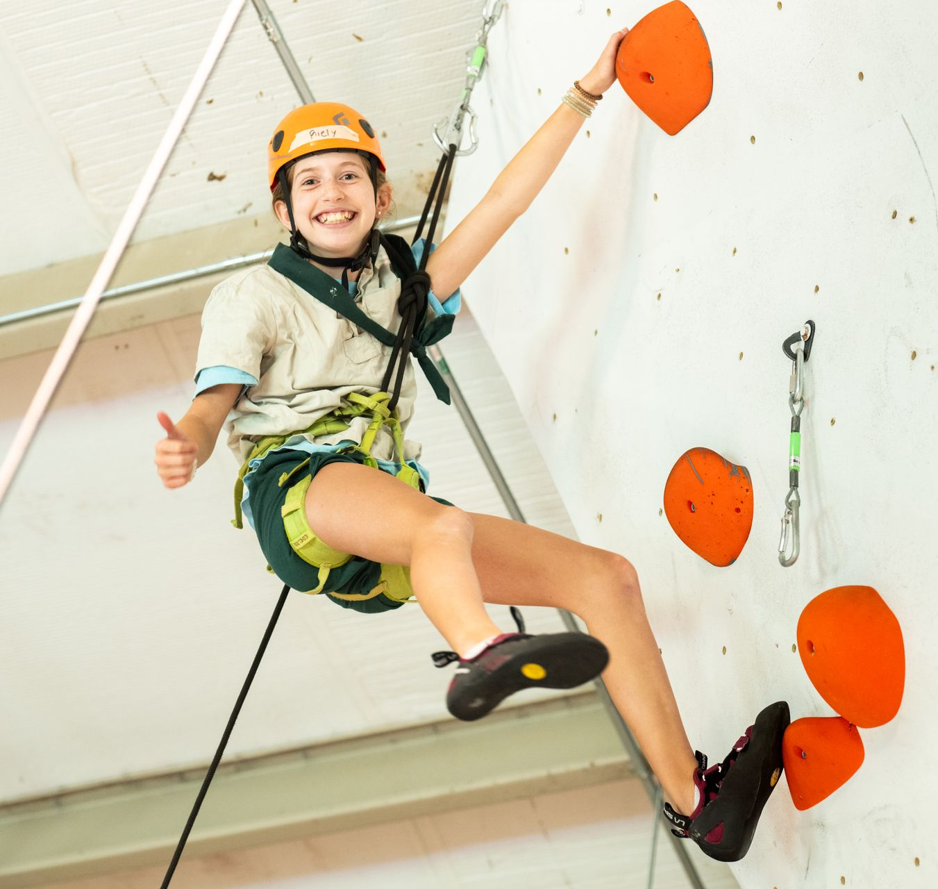 A smiling climber in a helmet and harness reaches for an orange hold on an indoor wall while giving a thumbs-up.