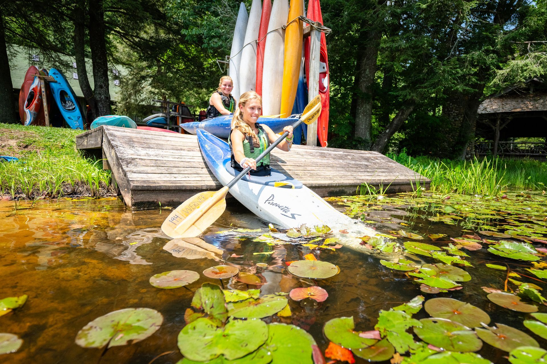 Two people in kayaks next to a wooden dock on a lake filled with lily pads, with a rack of colorful kayaks in the back.