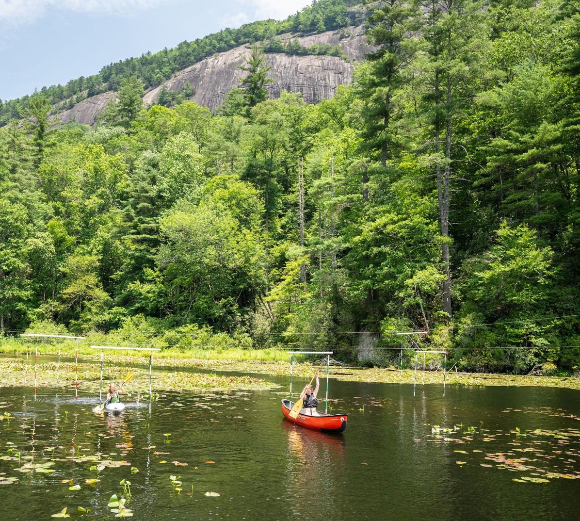 People paddling canoes on a lake filled with lily pads, framed by a dense forest and a large mountain rock face.
