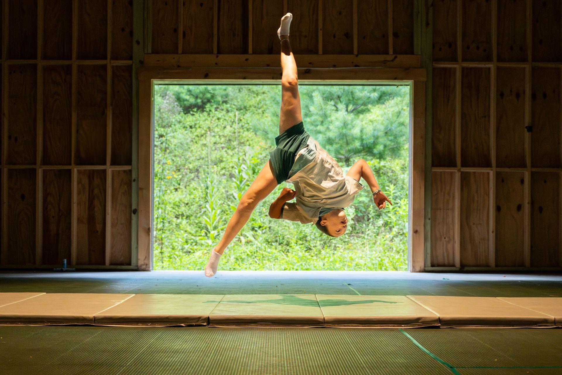 A person in athletic wear performs an aerial mid-air leap in a rustic, open-walled structure overlooking greenery.