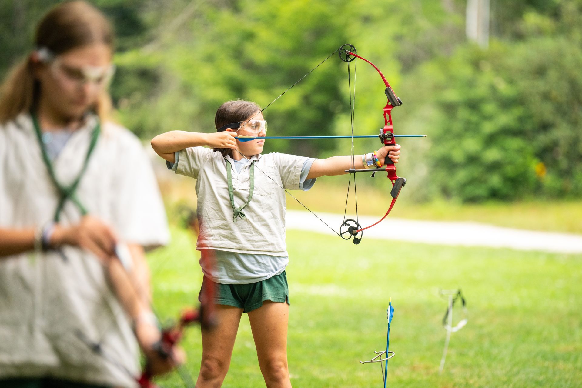 A person aims a compound bow at an outdoor archery range while another stands in the foreground, out of focus.