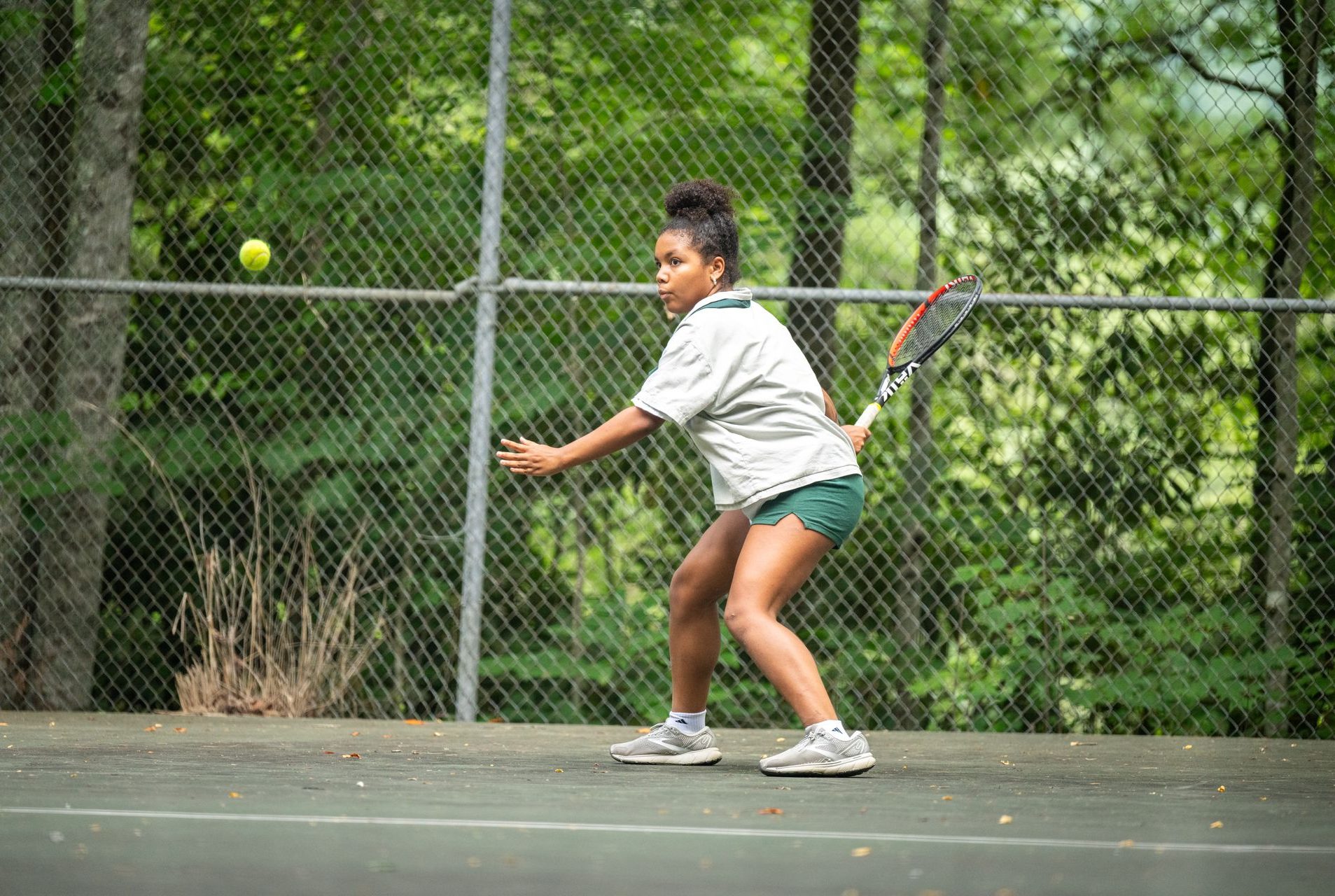 A person in a light-colored shirt and green shorts hits a tennis ball on an outdoor court with a chain-link fence behind.