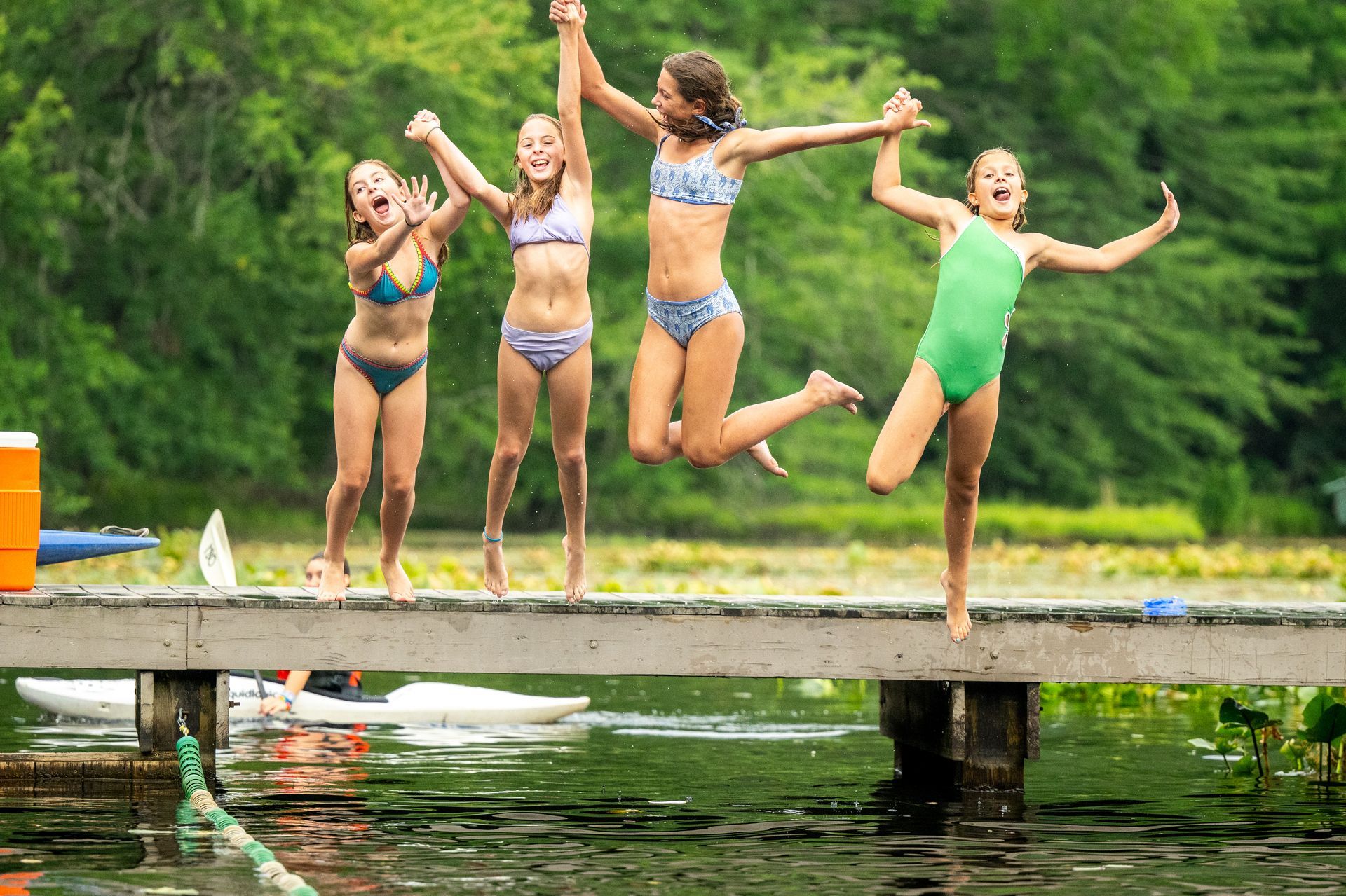 Four children jump joyfully from a wooden dock into a lake surrounded by green trees.