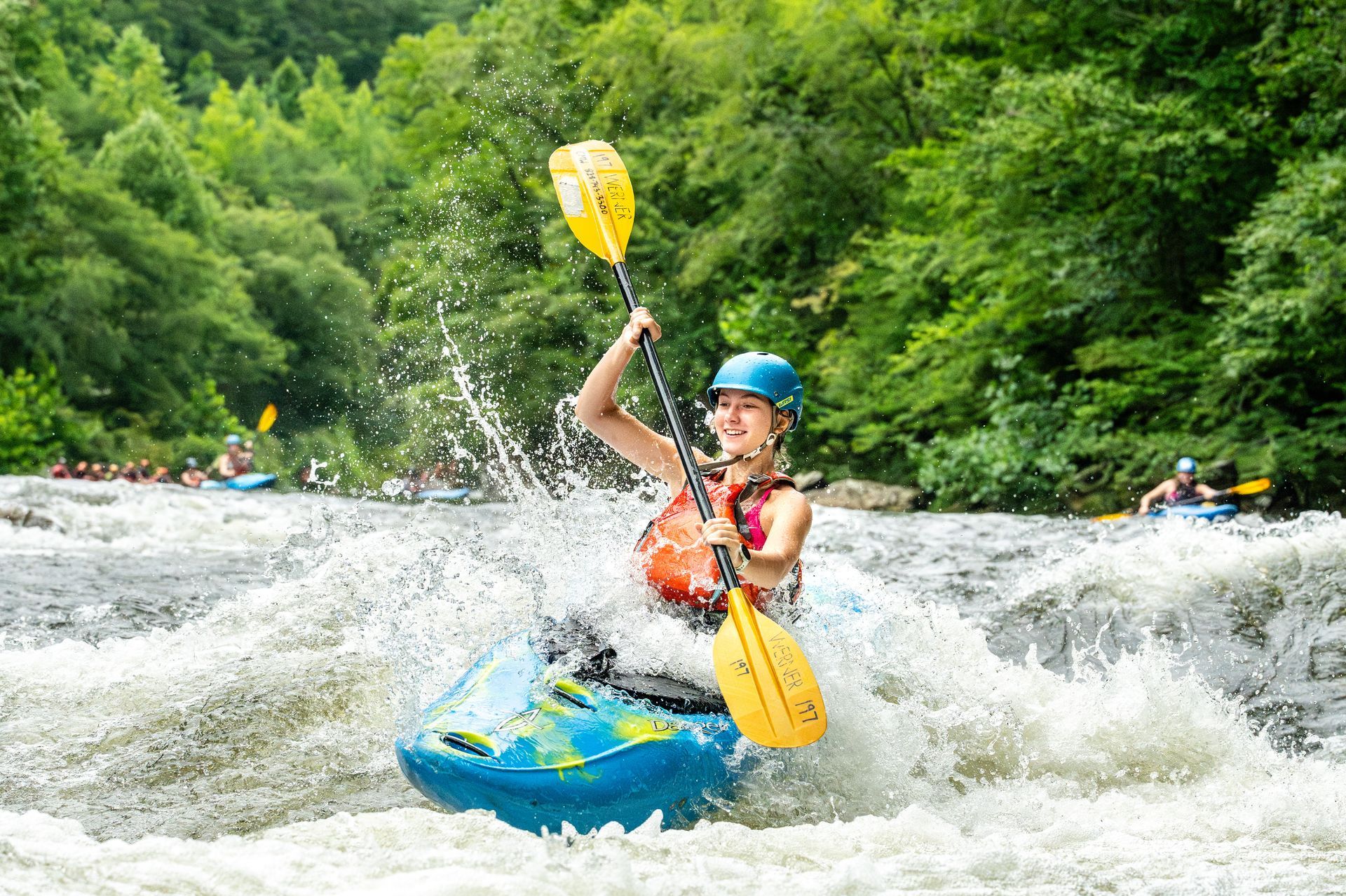 A person in a blue helmet and life vest paddles a bright blue kayak through splashing whitewater rapids in a forest.