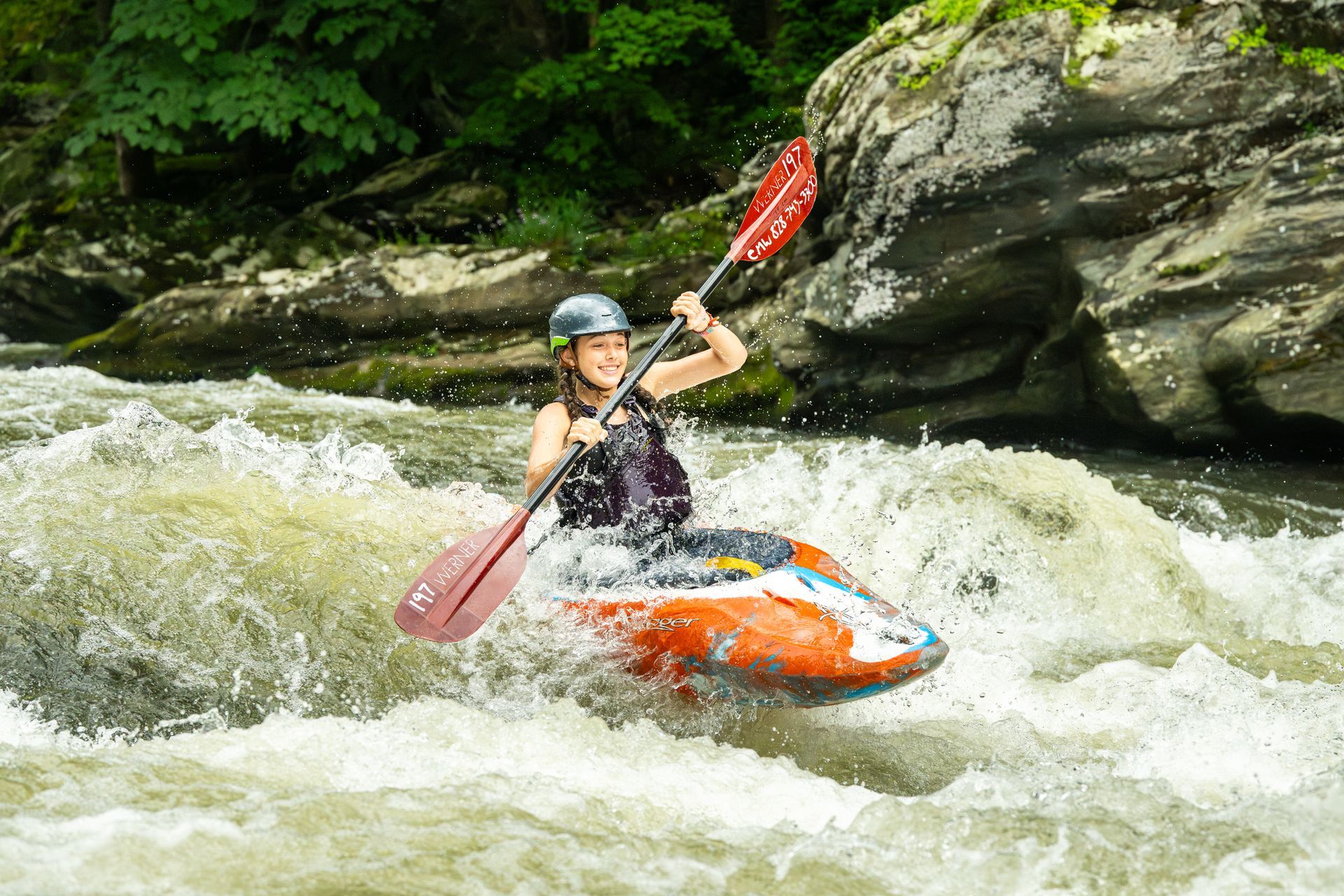 A person in a helmet and life vest whitewater kayaking down a river in an orange and white kayak.