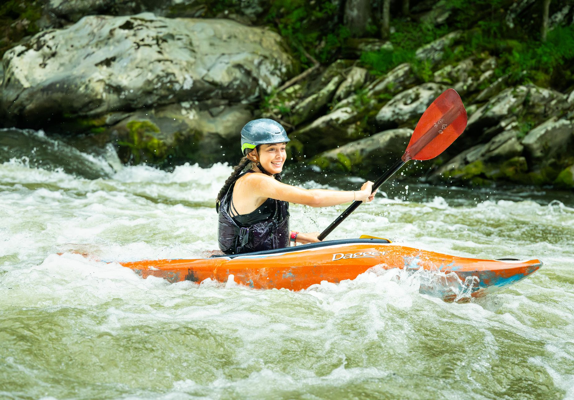 A person in a helmet and life vest smiles while kayaking in an orange kayak through choppy river rapids.