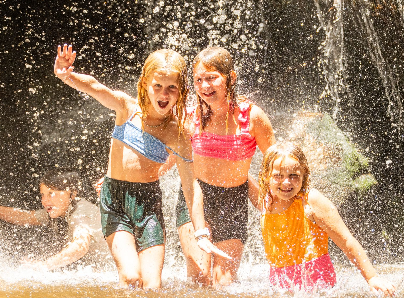 Four people splashing and smiling under a waterfall in bright swimwear.