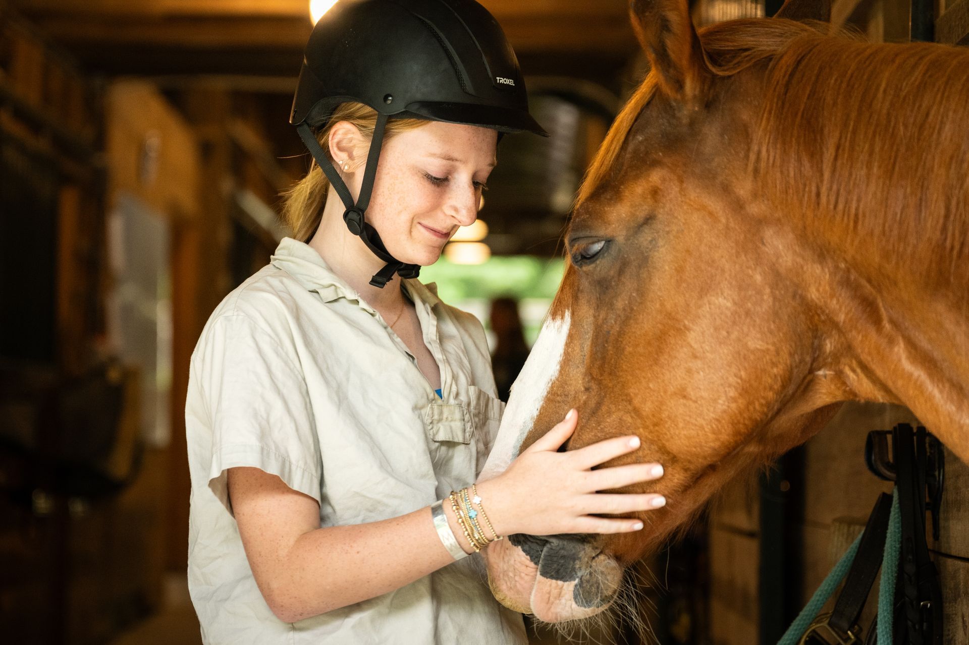 A person in a riding helmet gently pets a brown horse inside a stable.