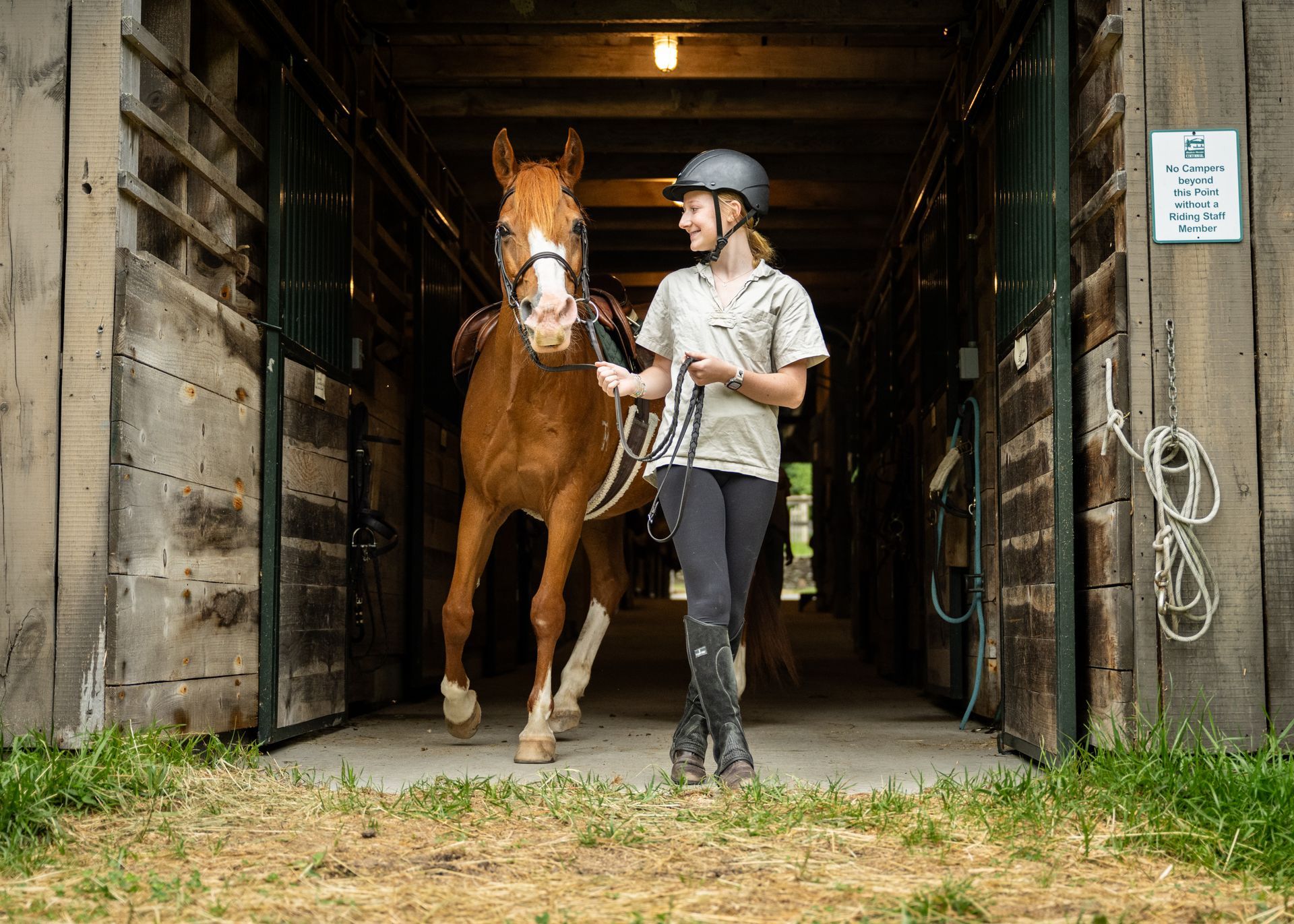 A person in riding gear leads a saddled chestnut horse out of a rustic wooden stable.