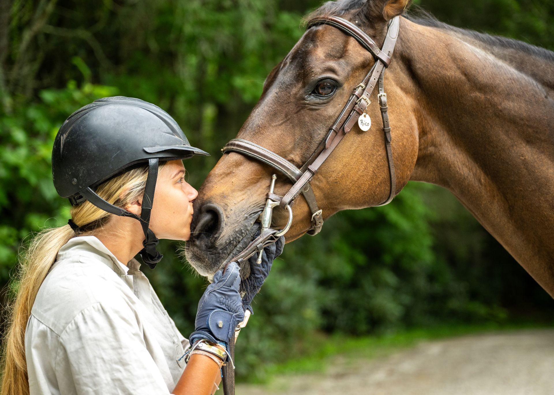 A person in a riding helmet and gloves kisses a brown horse on the nose, set against a blurred green, wooded background.