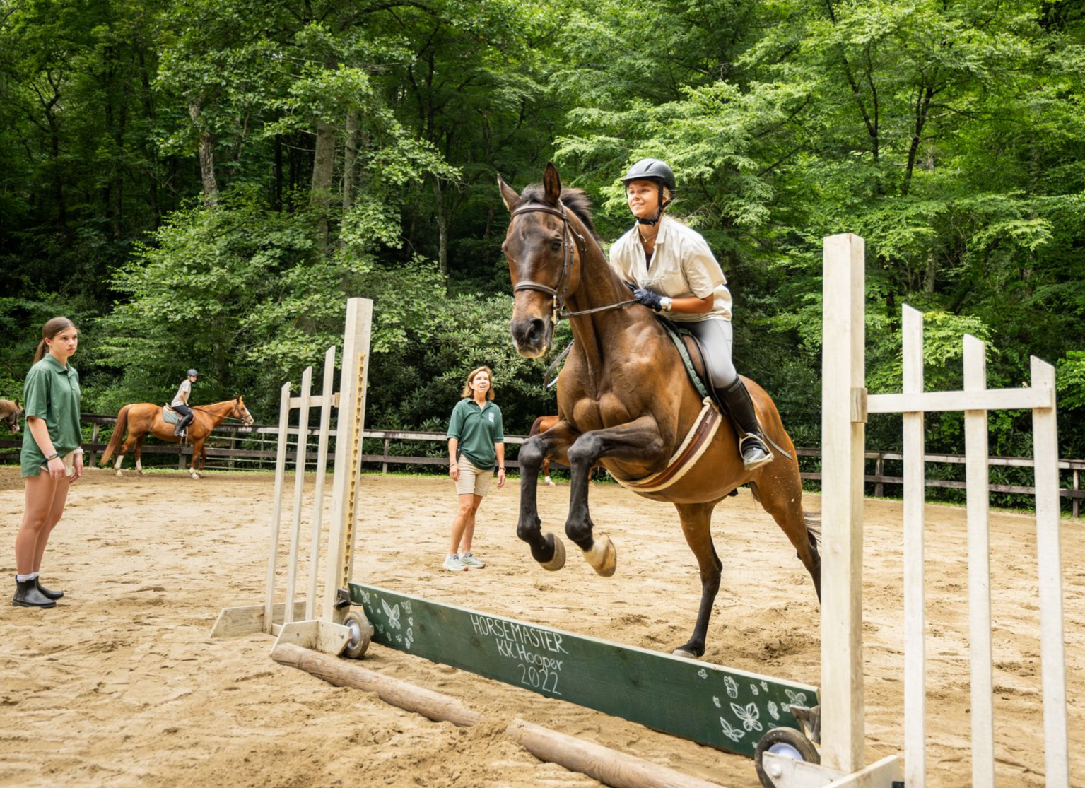 A person on a brown horse jumping over a wooden hurdle in a dirt riding ring with two people standing nearby.