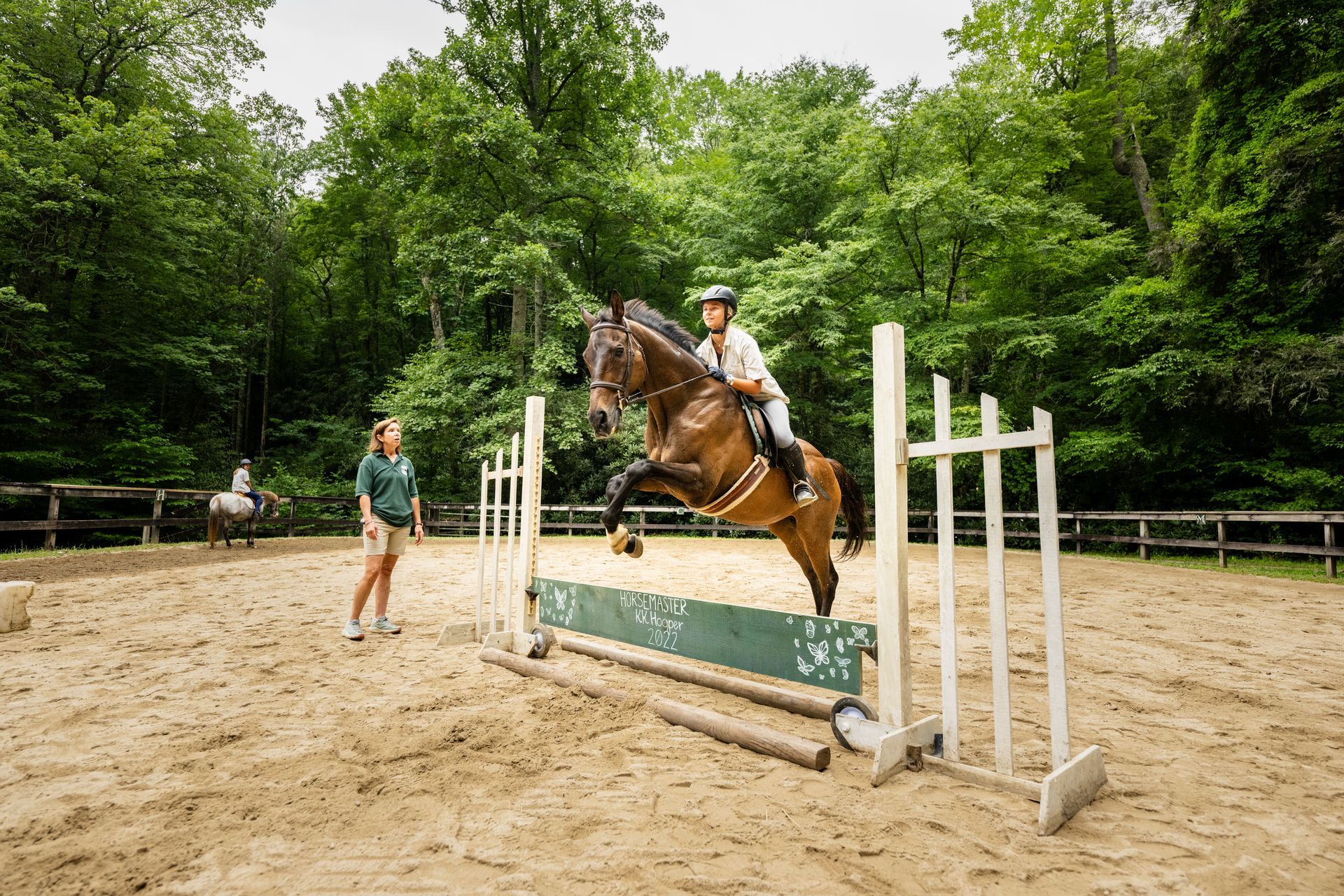 A rider on a brown horse jumps over a green obstacle in a sandy ring, while an instructor watches nearby.