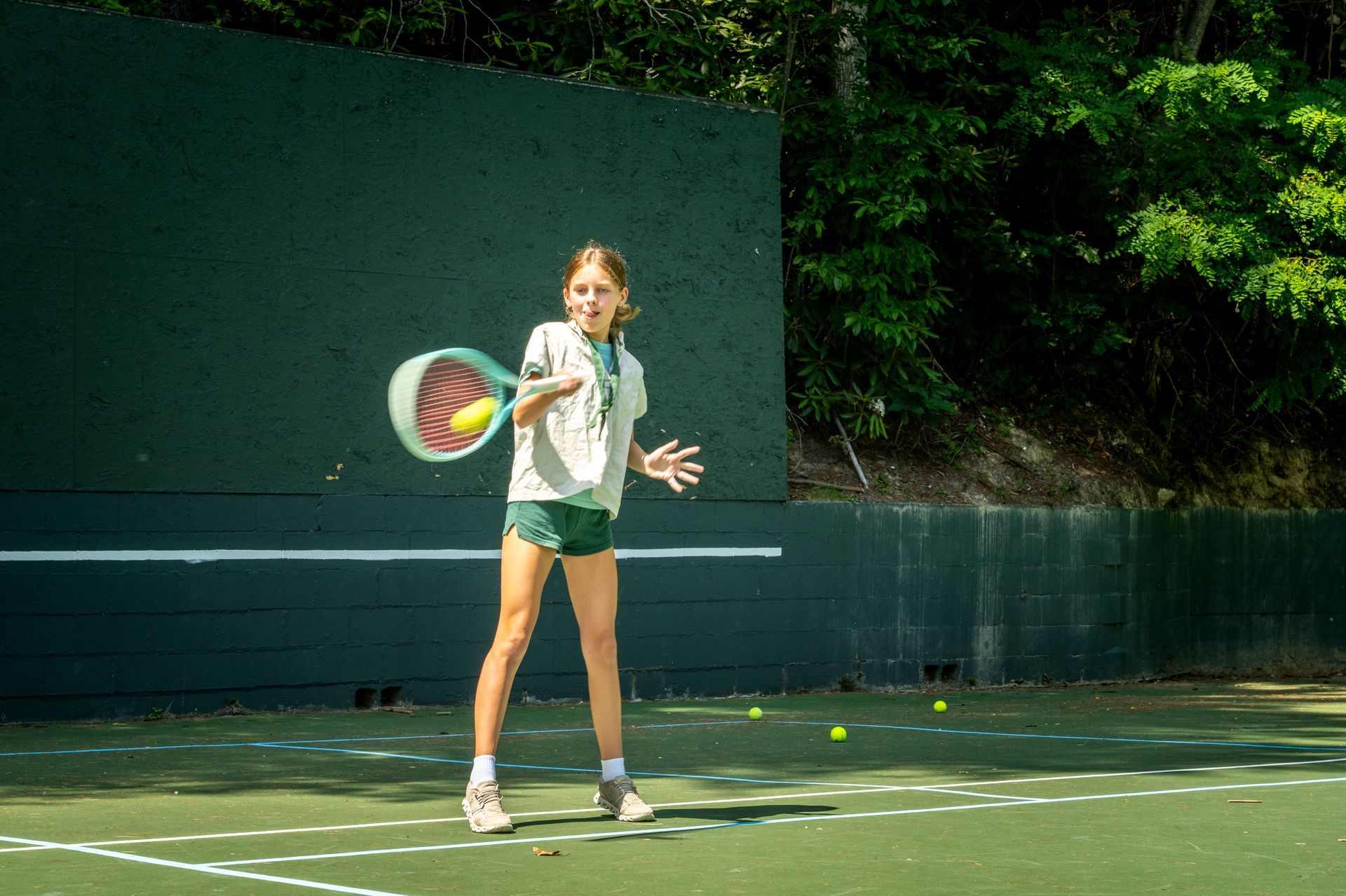 A person in athletic shorts and a t-shirt hits a tennis ball against a dark green practice wall on a court.