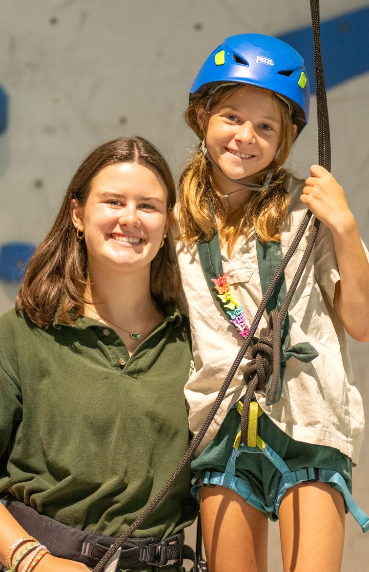 A person in a green polo shirt stands beside a smiling child in a blue climbing helmet and harness in front of a wall.