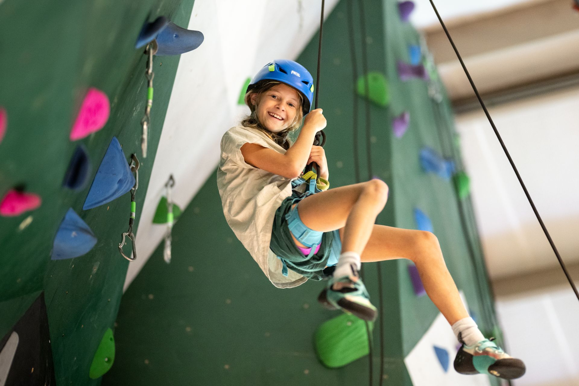 A person wearing a blue helmet and climbing harness smiles while suspended by a rope on an indoor rock climbing wall.