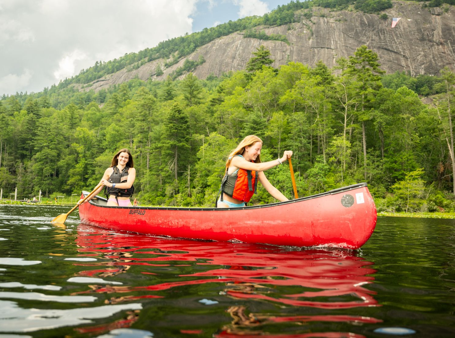 Two people paddle a red canoe on a calm lake, with green trees and a large rock mountain in the background.