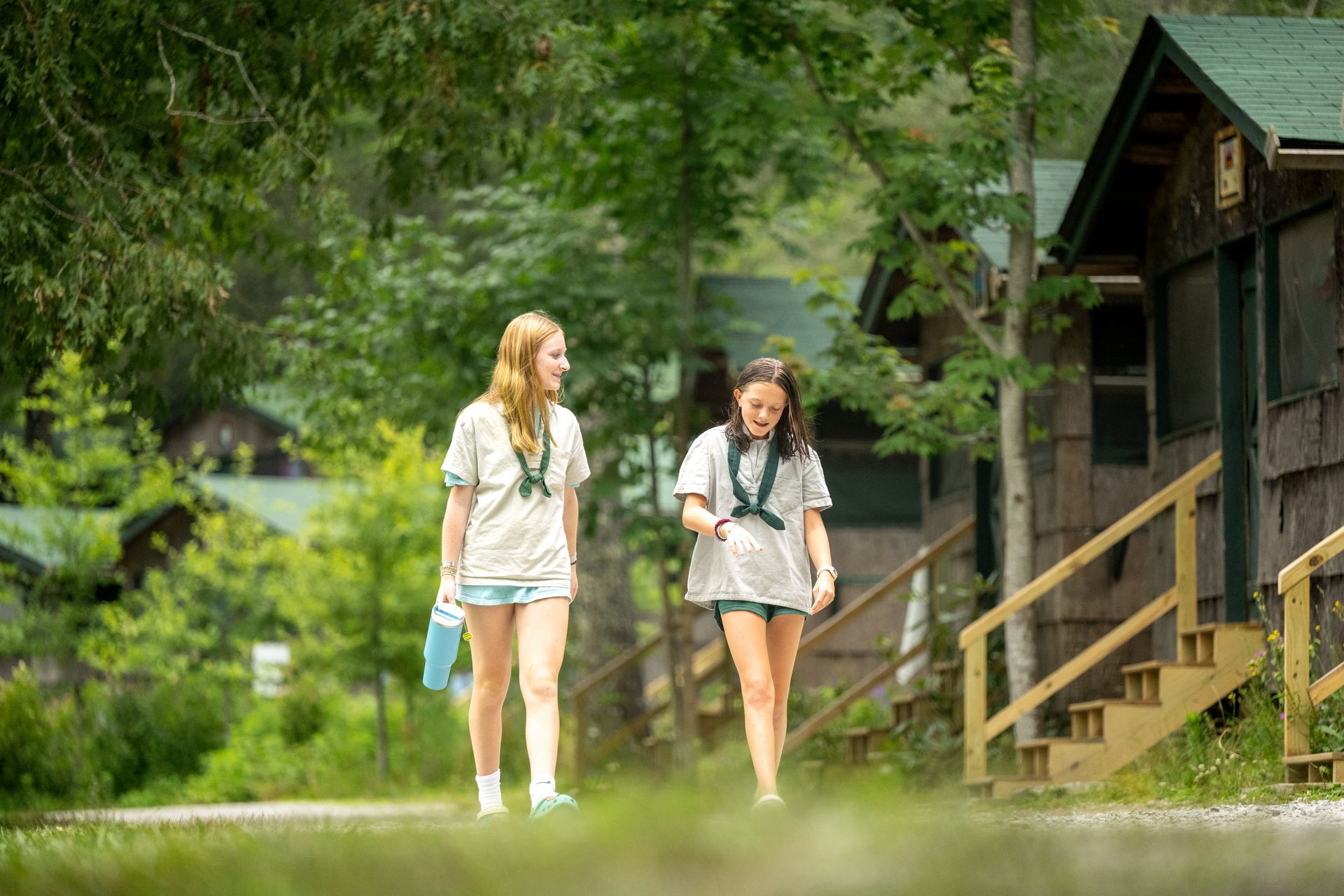 Two people in light-colored shirts and shorts walk and talk along a path past rustic cabins in a wooded area.