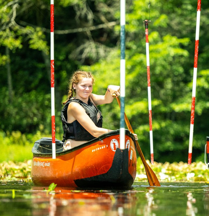 A person in a life jacket paddles an orange canoe through a slalom course of red and white poles on a water lily pond.
