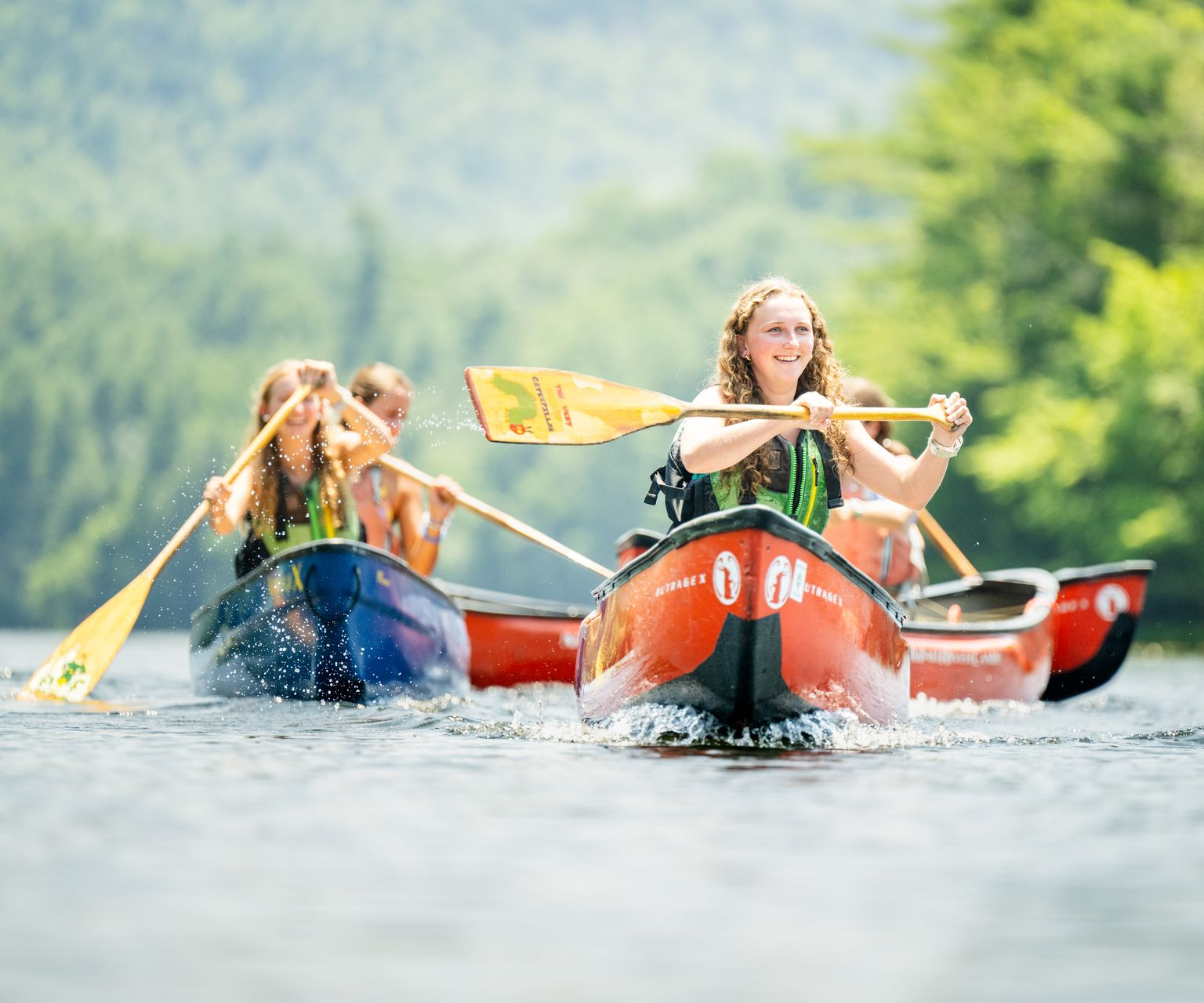 People paddling in red and blue canoes on a lake near a forest, with one individual in the foreground smiling.
