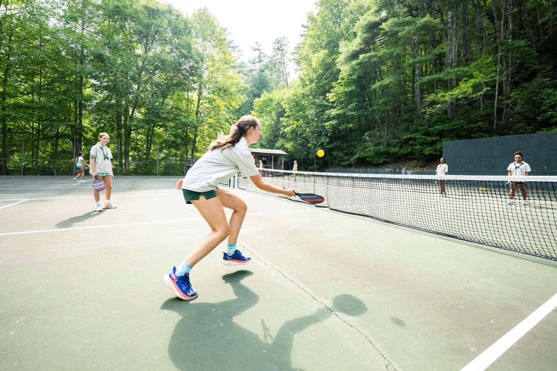 A person lunges to hit a yellow ball with a paddle on a pickleball court surrounded by trees.