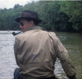 A person in a fedora, sunglasses, and tan jacket sits in a boat on a river, looking back while holding a pipe.