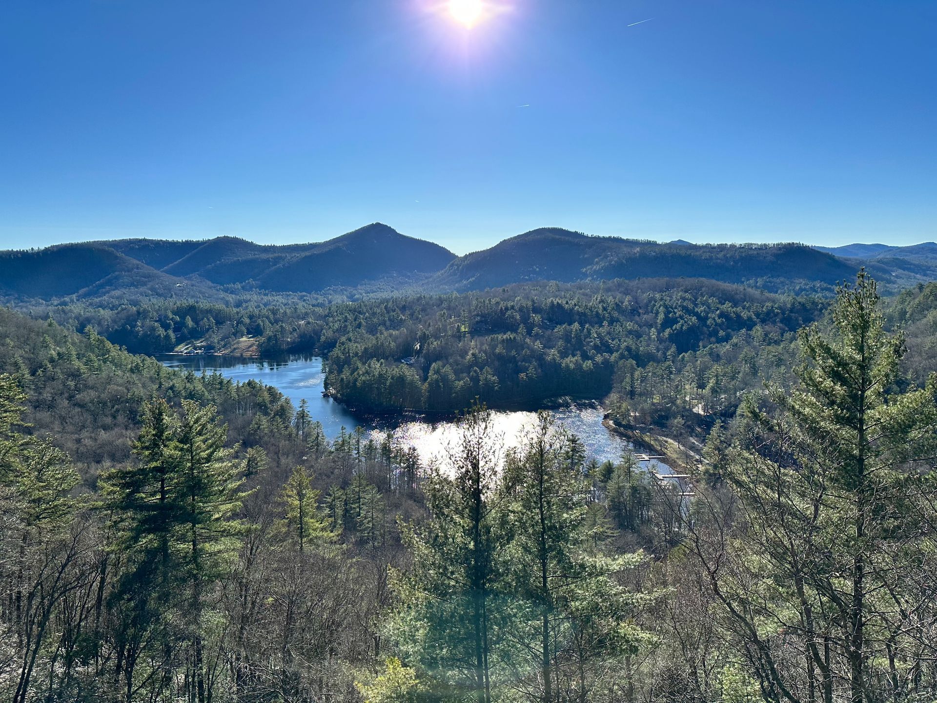 A bright sun shines over a tranquil lake surrounded by a dense, sunlit forest and distant rolling mountain ridges.