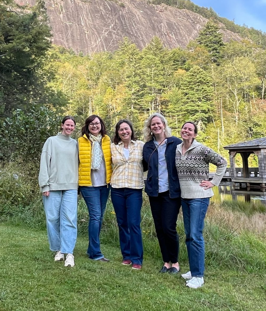 Five people stand in a line on a grassy field before a forested mountain and a lakeside structure, smiling at the camera.